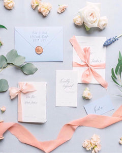 image of a pastel flat lay photo on a pale gray background with pink ribbon, calligraphy invitation suite, broze wax seal and flowers.